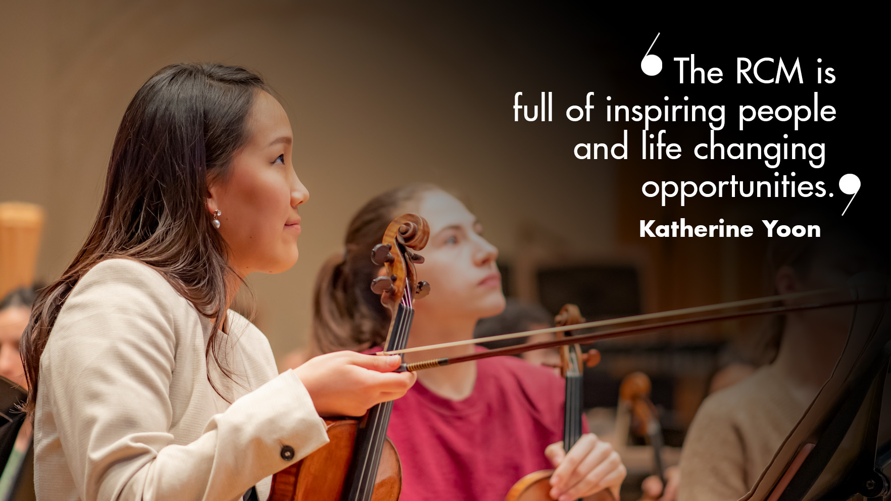 “The RCM is an incredible place, full of inspiring people and life changing opportunities". A female student, wearing a white jacket, sitting in an orchestral rehearsal, holding her bow and violin.